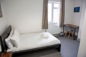 Una cama individual con ropa de cama blanca, cabecera de madera y una toalla enrollada. Hay un escritorio pequeño con una silla, un radiador y una ventana con cortinas.
