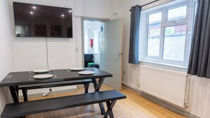 Ruokailutila, jossa on neljälle katettu pöytä, penkki, seinälle asennettu taulutelevisio ja ikkuna, jossa on verhot.