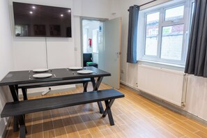 Uma área de refeições com mesa posta para quatro, um banco, uma TV de tela plana na parede e uma janela com cortinas.