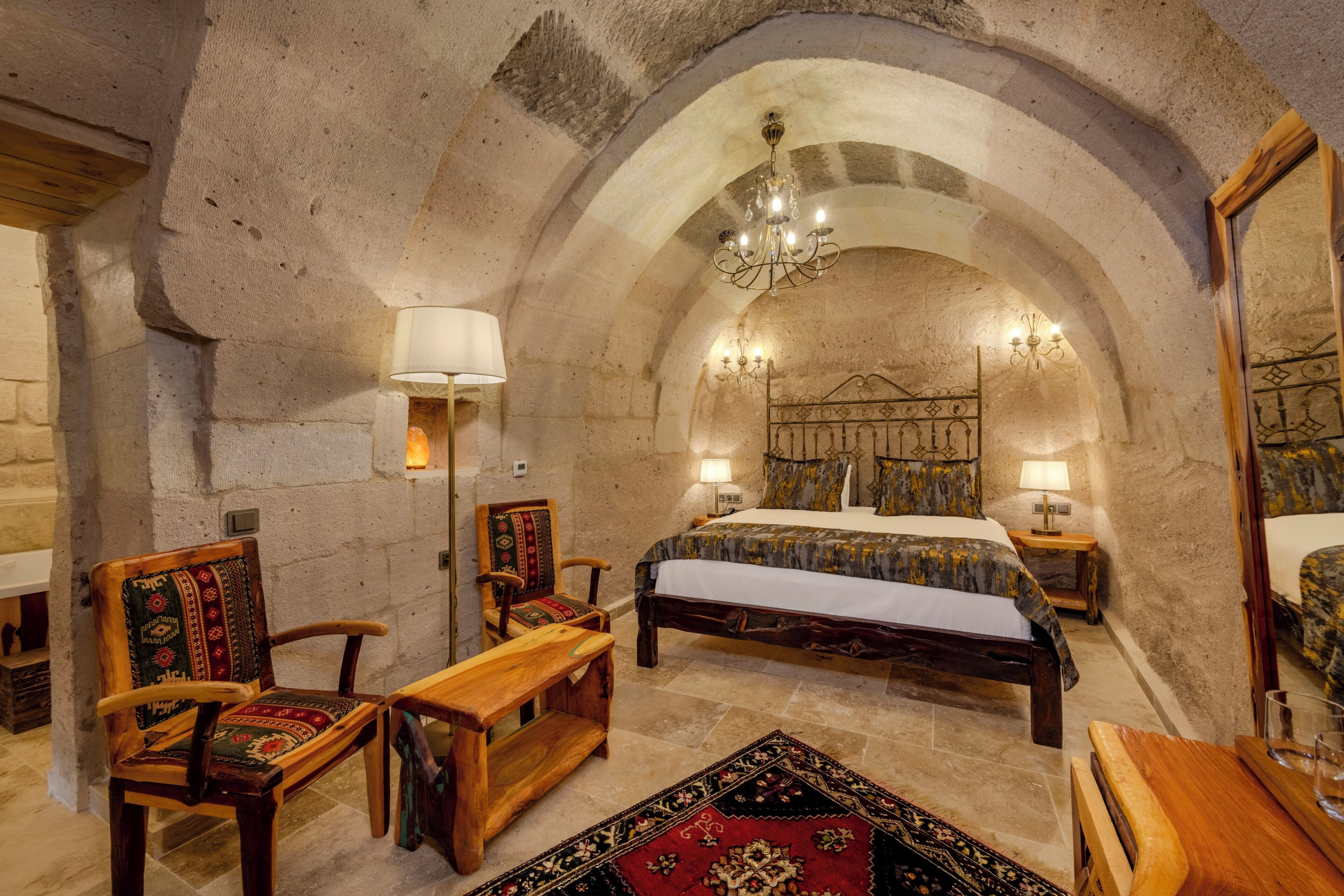 A room with a vaulted ceiling, a bed with a patterned bedspread, two chairs with cushions, a wooden bench, and a chandelier.
