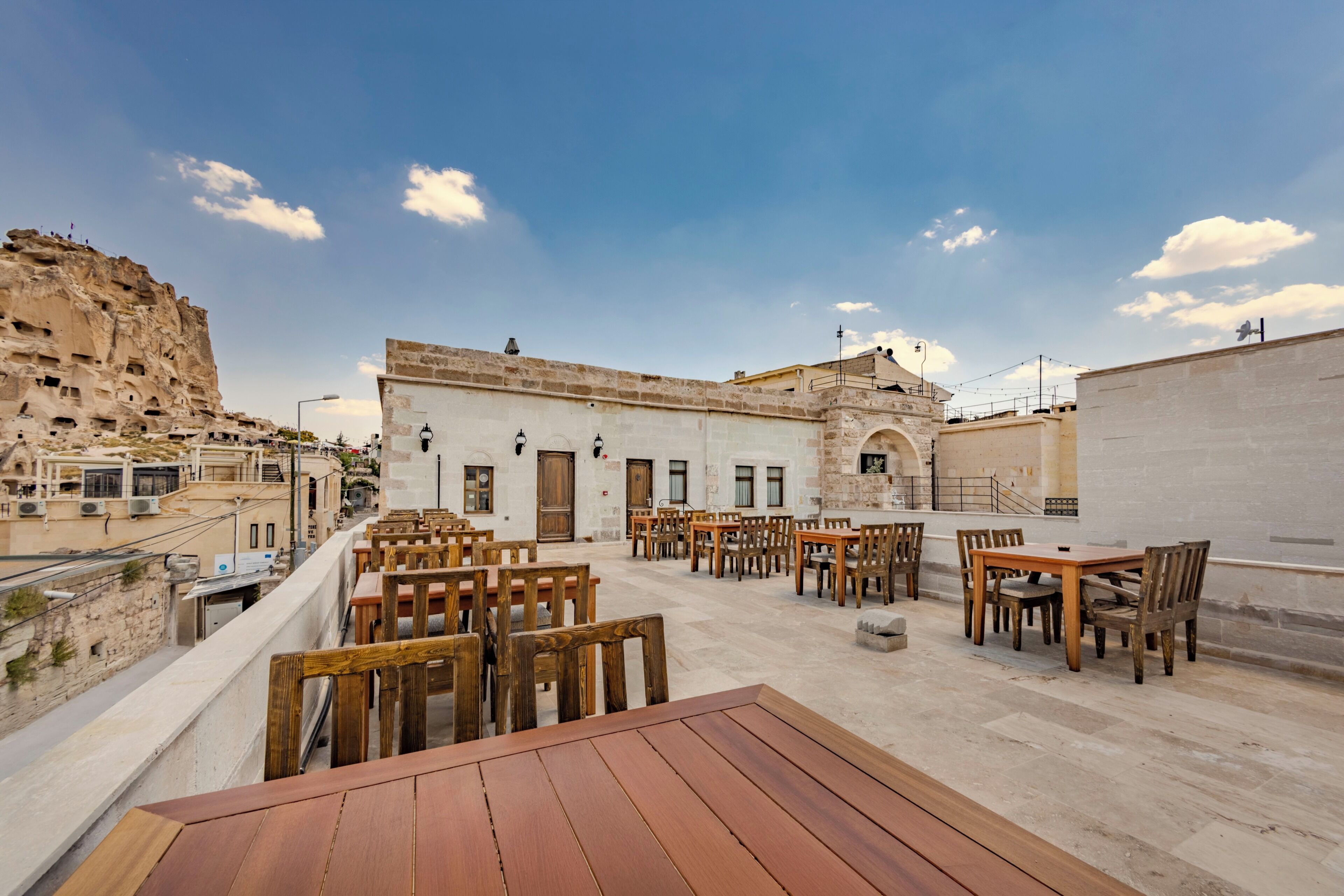 Une terrasse sur le toit, avec des tables et des chaises en bois, un bâtiment en pierre en arrière-plan et un ciel dégagé.