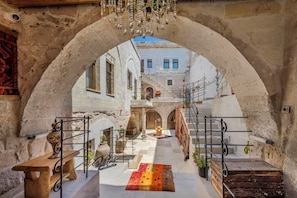 Un patio interior con un arco de piedra, un banco de madera, una mesa, plantas en macetas y una escalera que conduce a un nivel superior.