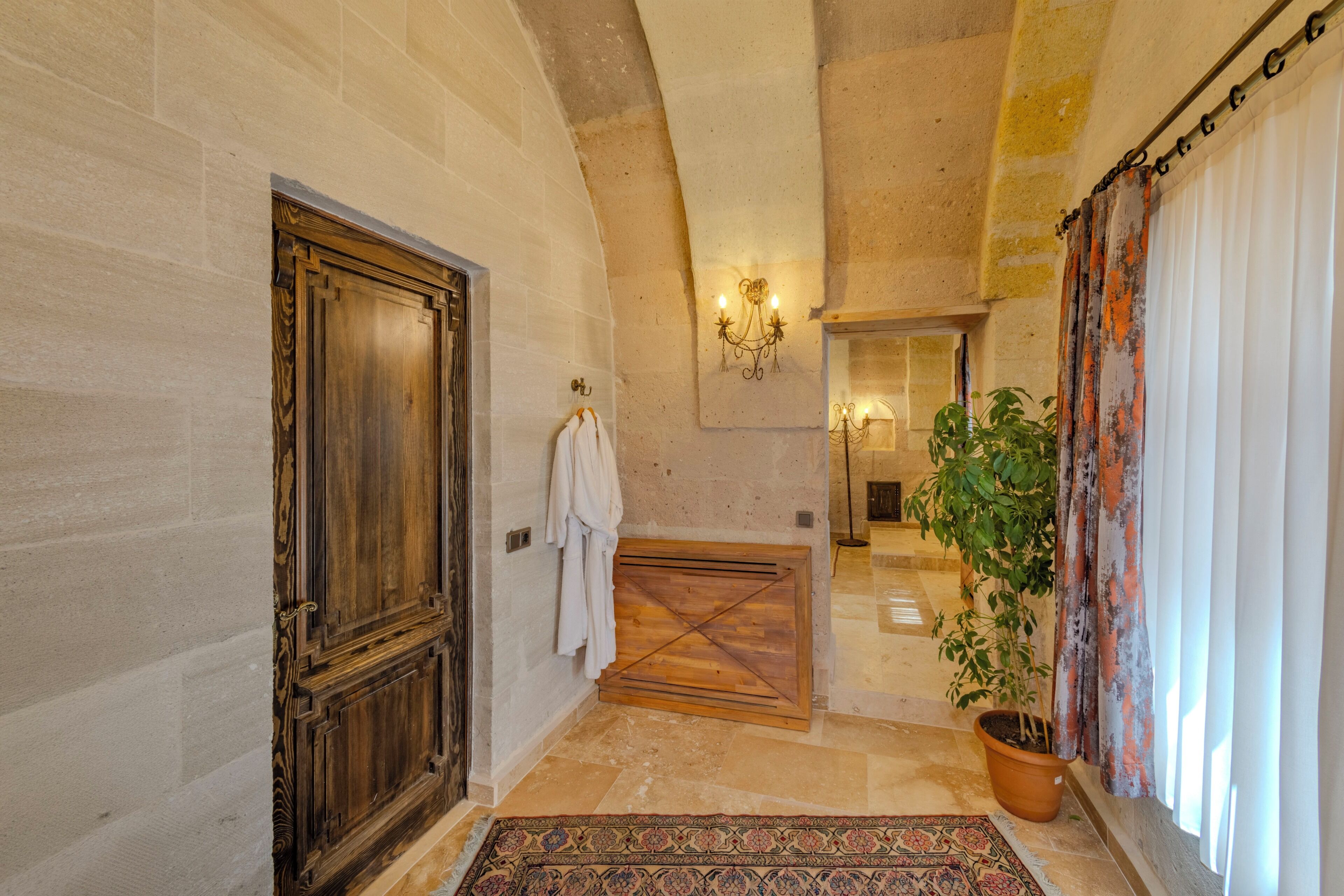 A hallway with a wooden door, a white robe hanging on the wall, a potted plant, and a patterned rug.