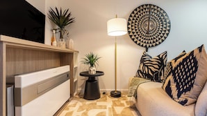 A modern living room with a flat-screen TV, a sofa with patterned cushions, a floor lamp, and a decorative wall piece. - Neo-African studio in Cape Town with spectacular rooftop swimming views (Cape Town)