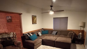 Una sala de estar con un sofá modular, un sofá más pequeño, una alfombra, un ventilador de techo, un cuadro enmarcado y una ventana con persianas.