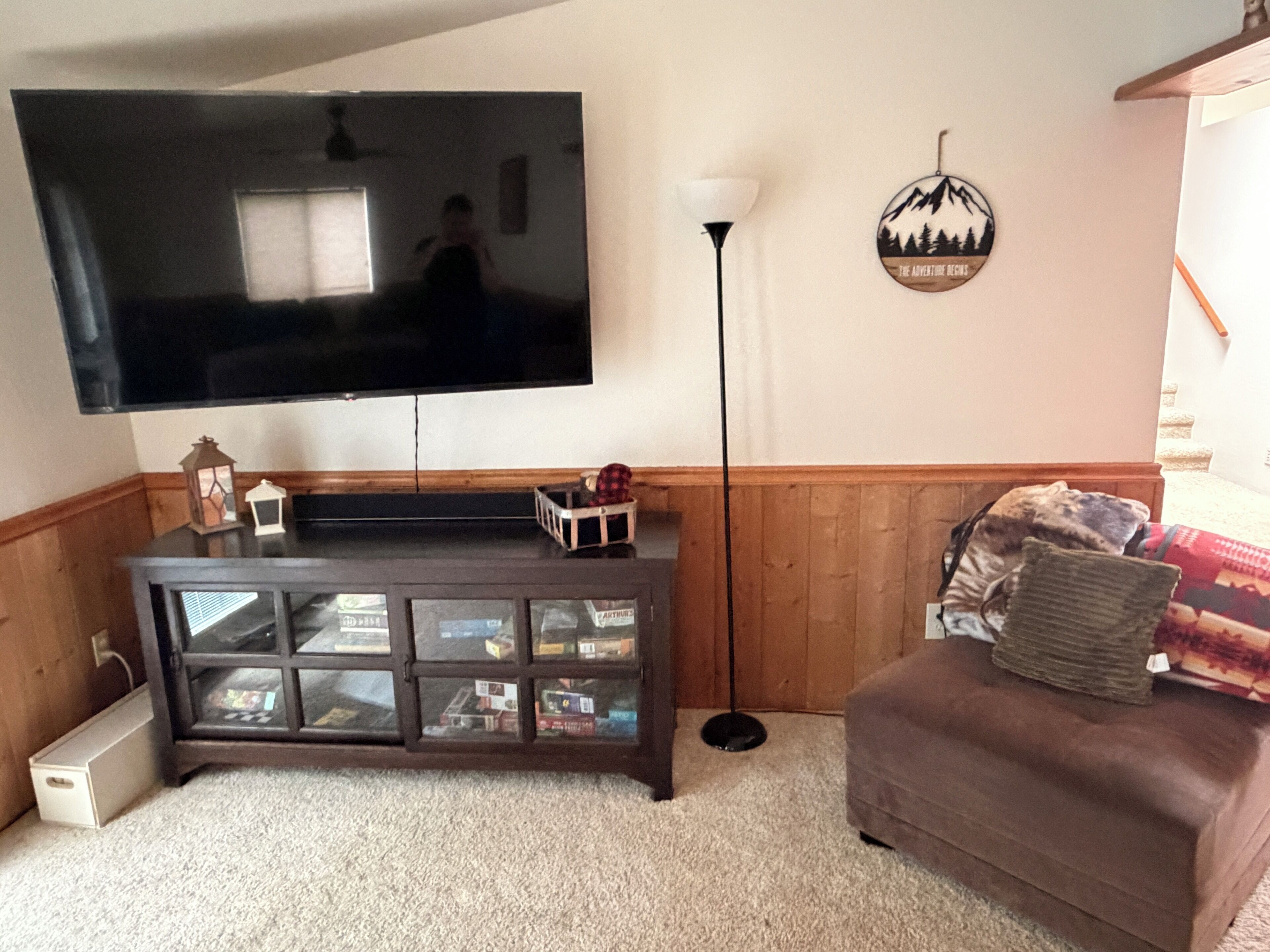 A living room with a flat-screen TV mounted on the wall, a dark wooden entertainment center, a floor lamp, and a brown sofa with patterned cushions.