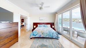 Ein Hotelzimmer mit einem Bett, einer Holzkommode, einem Deckenventilator, einem großen Fenster mit Meerblick und einer Essecke im Hintergrund.
