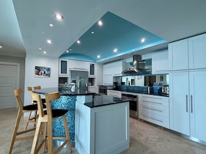 Eine moderne Küche mit weißen Schränken, einer schwarzen Arbeitsplatte und einer blauen Spritzschutzwand. Zwei Holzstühle und ein Esstisch sind vorhanden.
