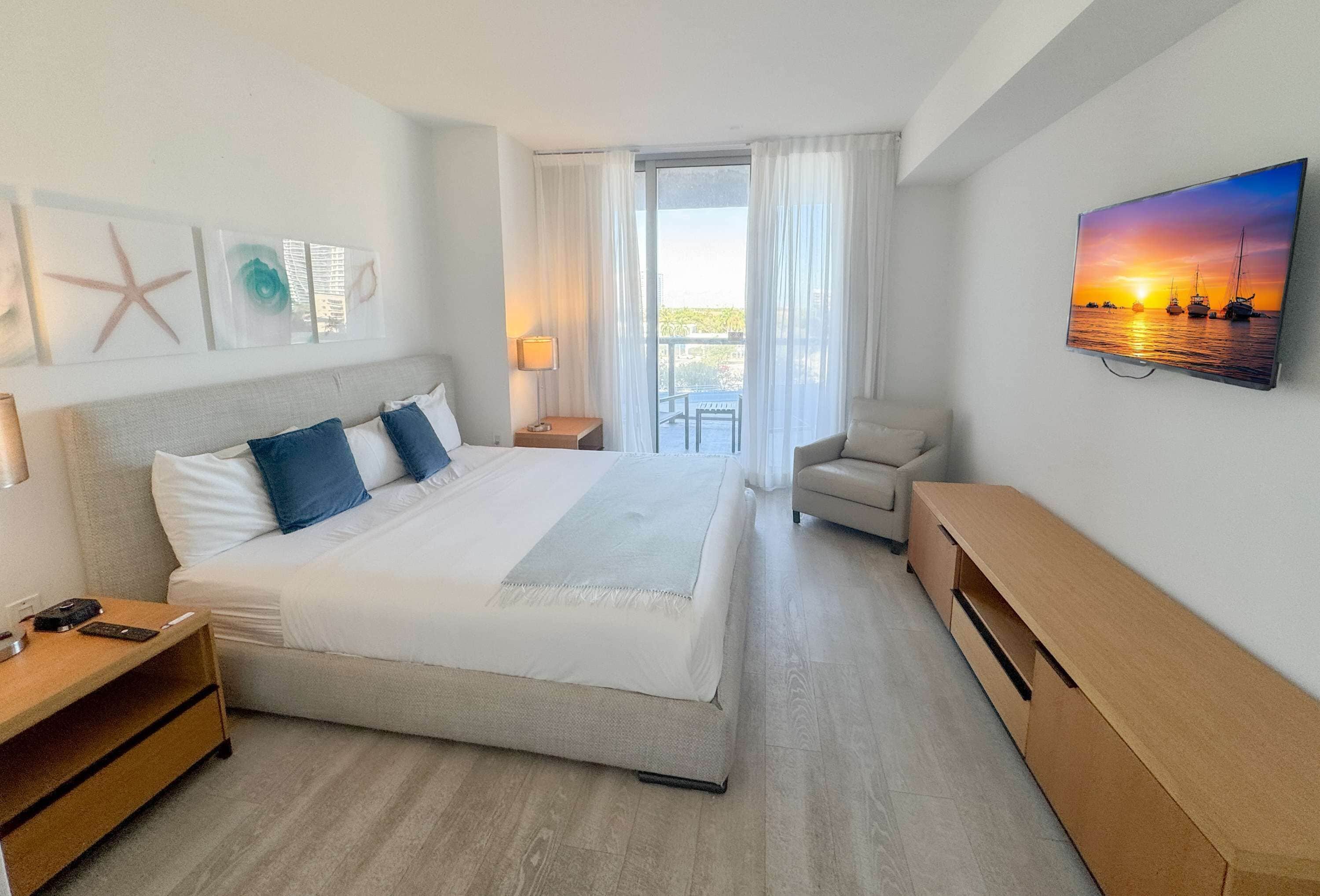 Quarto de hotel com uma cama grande, uma mesa, uma cadeira, uma televisão e varanda com vista para o mar.