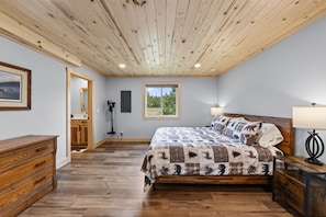 Une chambre avec un plafond en bois, un grand lit avec une literie à motifs, une table de chevet, une commode et une fenêtre donnant sur des arbres.