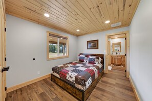 A bedroom with a wooden ceiling, a large bed with a patterned quilt, a window, and a bathroom with wooden cabinets. - Yellowstone Retreat w/AC & wrap-around porch. Fly fish, trail riding, snowmobile (Island Park)