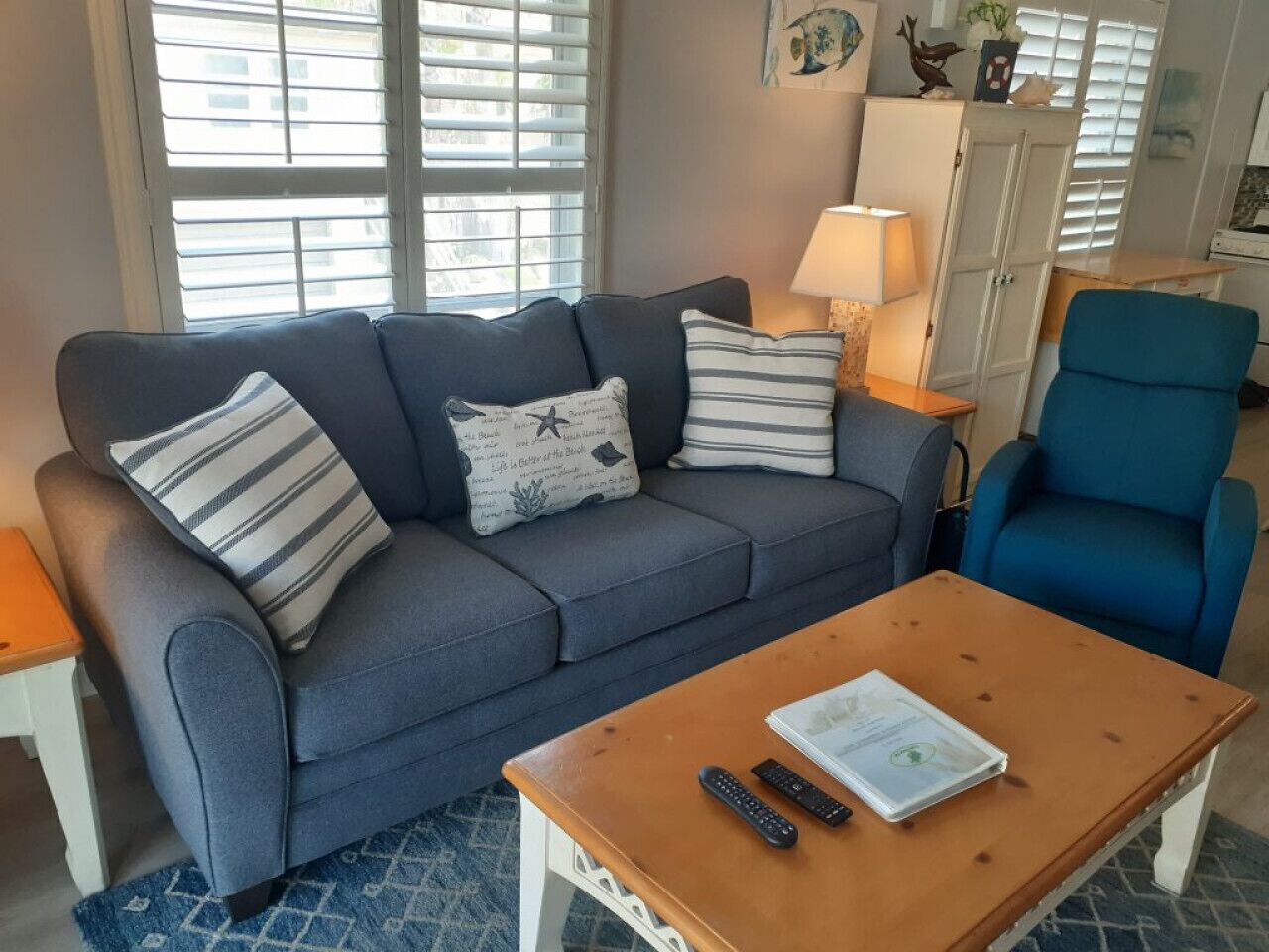 A living room with a grey sofa, striped pillows, a blue armchair, a wooden coffee table with a remote control and a booklet, and a lamp on a side table.