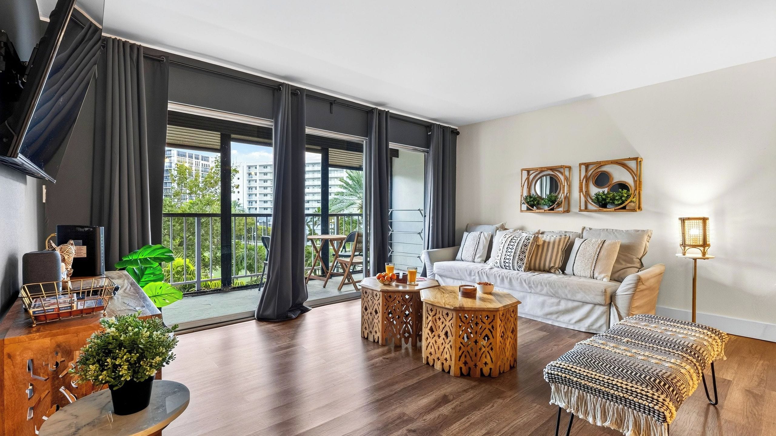 A modern living room with a sofa, coffee table, and side tables. There is a balcony with a view of buildings and greenery.