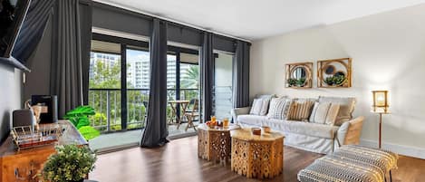 A modern living room with a sofa, coffee table, and side tables. There is a balcony with a view of buildings and greenery.