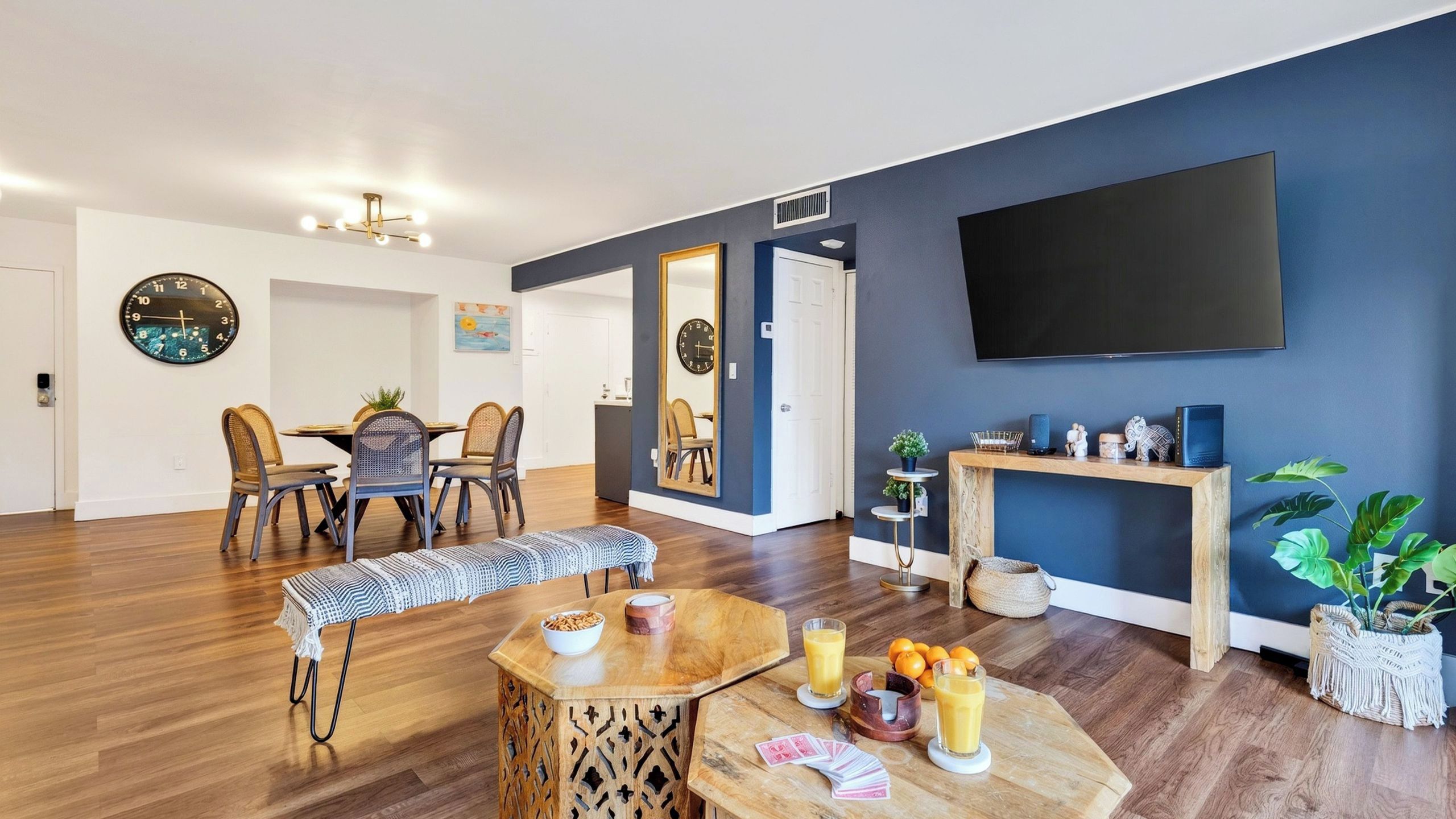 A modern living room with a dining area, wooden flooring, a flat-screen TV, and a large mirror.