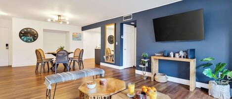 A modern living room with a dining area, wooden flooring, a flat-screen TV, and a large mirror.
