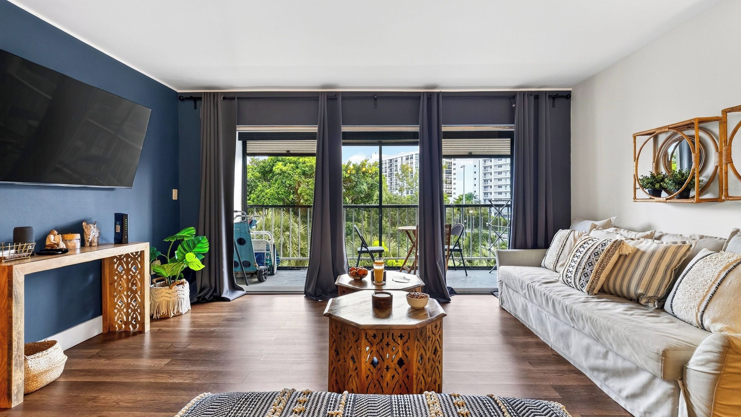 A modern living room with a large TV, a sofa, a coffee table, and a balcony with a view of greenery and buildings.