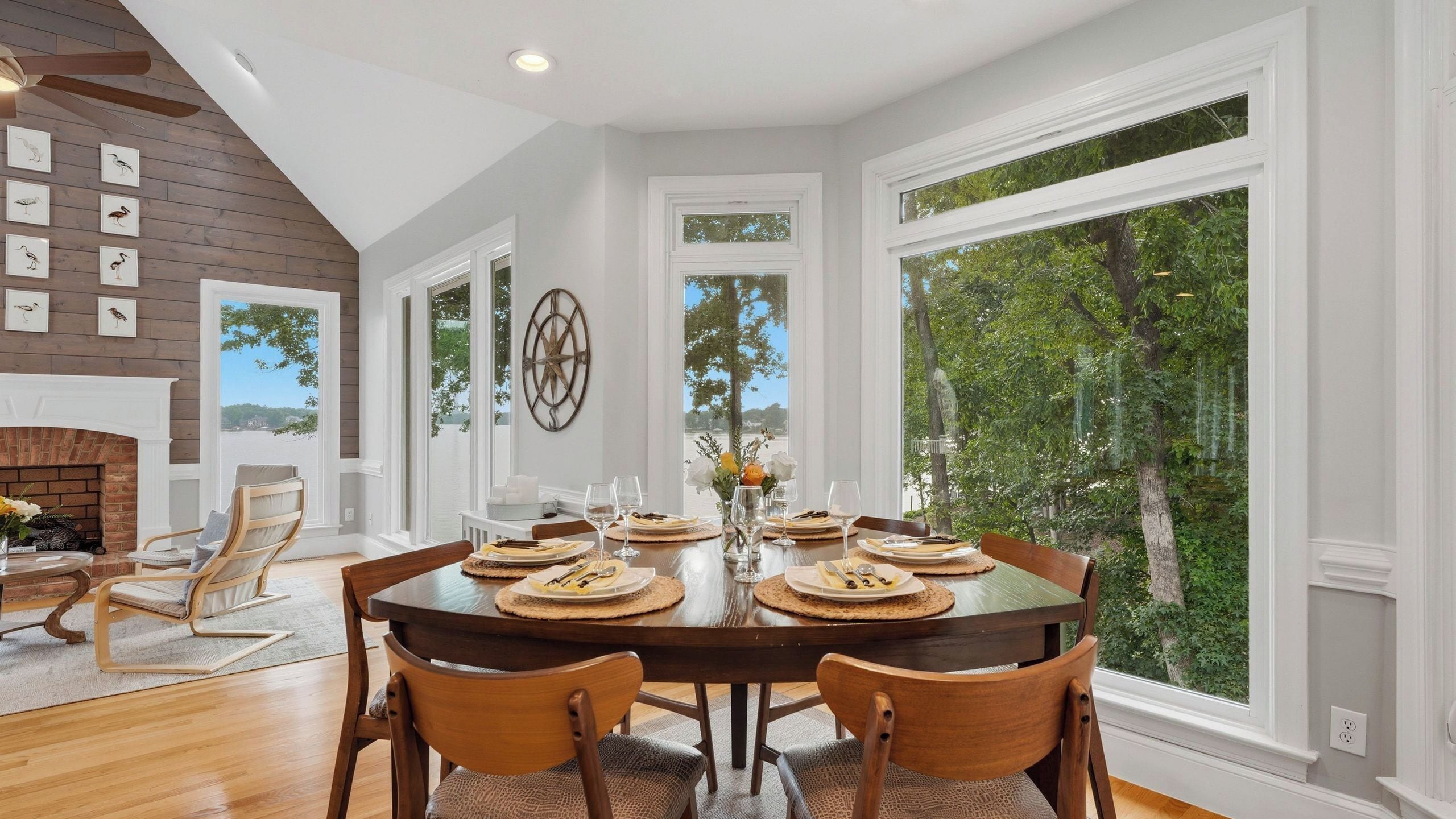 A dining area with a large table set for six, surrounded by wooden chairs, with a view of trees through large windows.