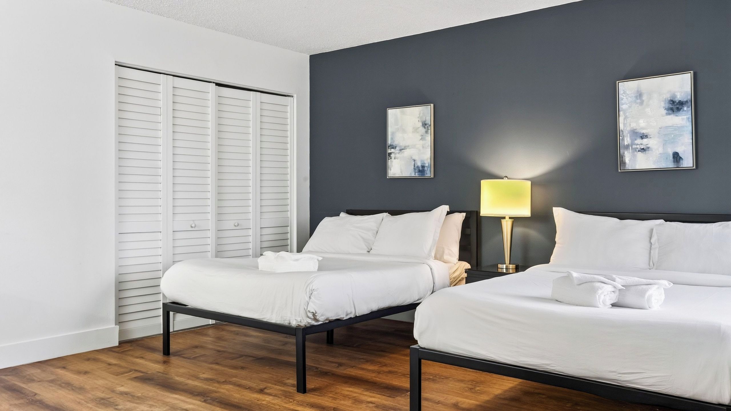 Une chambre d’hôtel avec deux lits, un plancher en bois, une armoire blanche et deux tableaux abstraits accrochés au mur.
