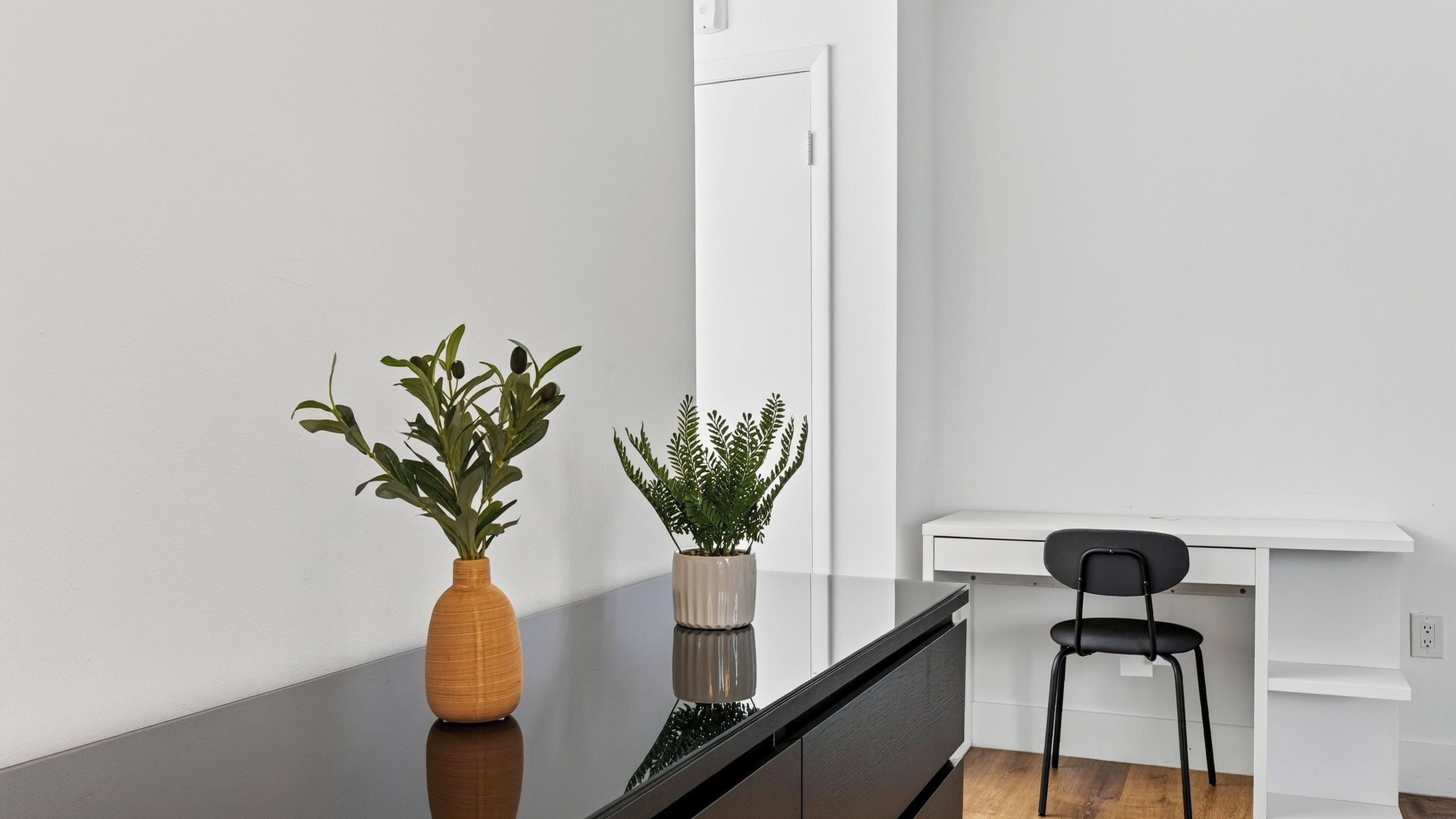 A minimalist room with a wooden table, two potted plants, a black chair, and a white wall.