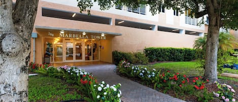 A hotel entrance with a paved pathway, a well-maintained garden with various flowers, and a building facade with the name 'MARBELLA TOWER'.