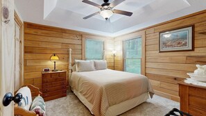 Ein Schlafzimmer mit einem Bett, Holzwänden, einem Deckenventilator, einem Nachttisch, einer Lampe und einem gerahmten Bild an der Wand.