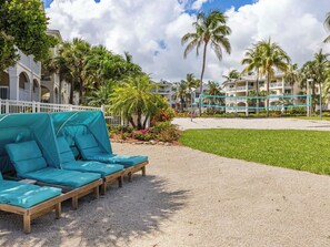 A beachside lounge area with a row of wooden lounge chairs and a covered seating area, surrounded by palm trees and a building with balconies. - Spacious Oceanside 2BR 2BA Condo w/Heated Infinity Pool - Hyatt Windward Pointe (Key West)