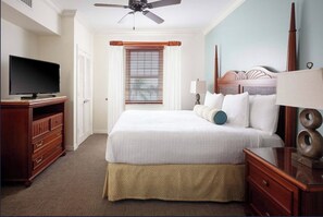 A hotel room with a bed, a television, a ceiling fan, a window with blinds, and a nightstand with a lamp.