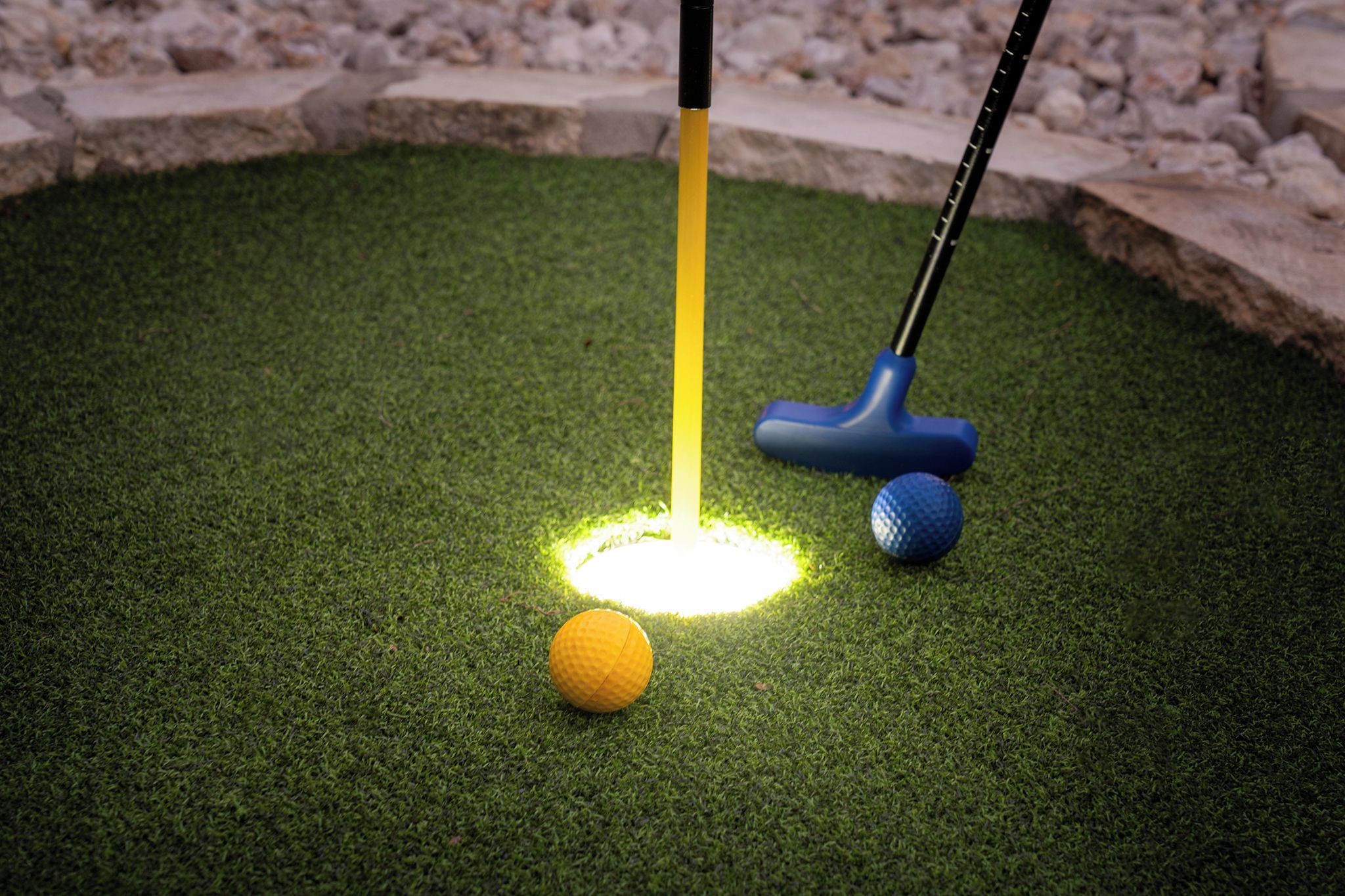 A miniature golf hole with a yellow ball and a blue ball on artificial grass, a golf club with a yellow shaft, and a blue club head.