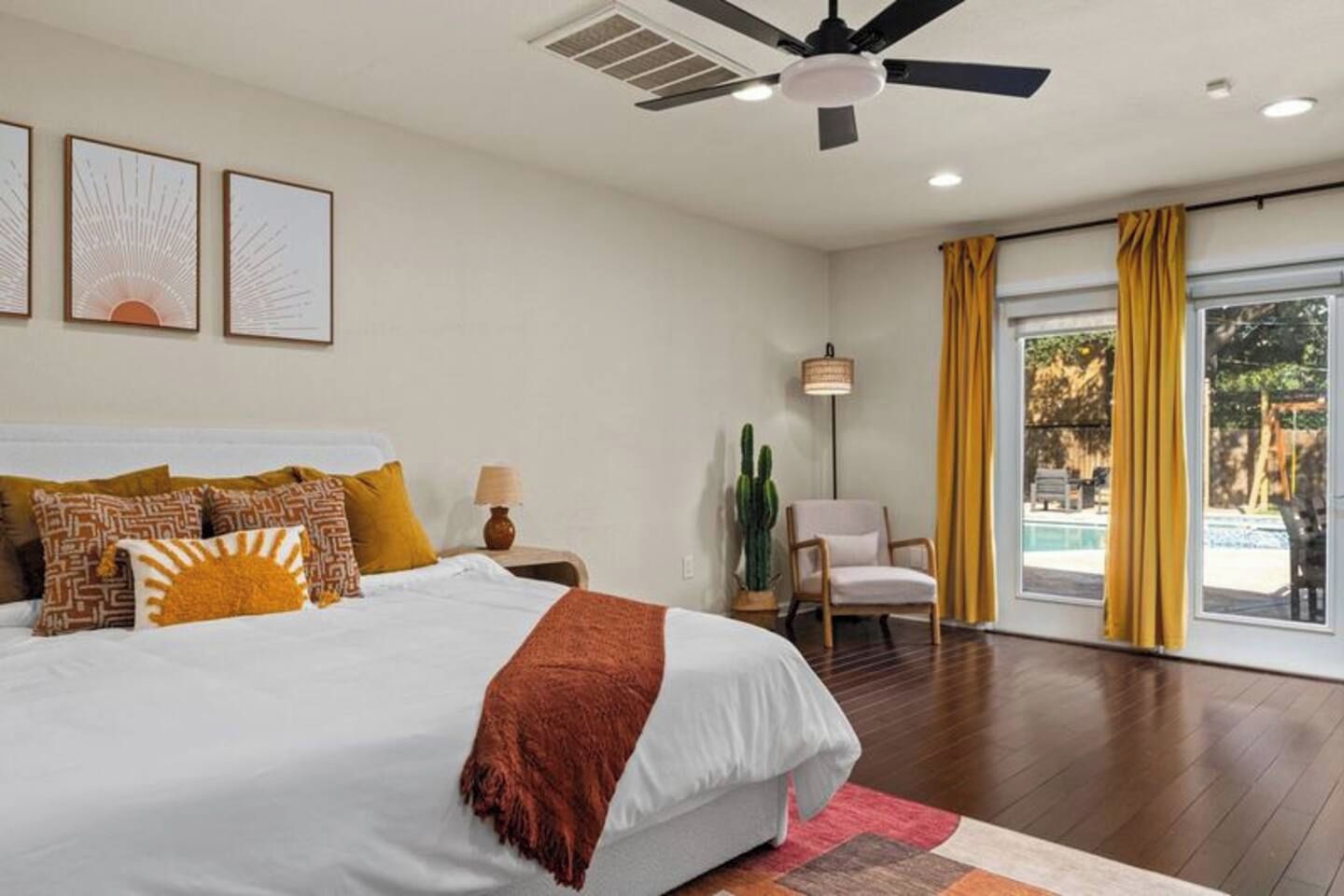 A bedroom with a large bed, a ceiling fan, a nightstand, a lamp, a chair, a cactus, and a view of an outdoor area.