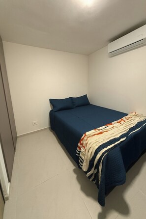 Ein kleines Zimmer mit einem Einzelbett, einer blauen Bettdecke, einer weißen Wand und einer unter der Decke angebrachten Klimaanlage.