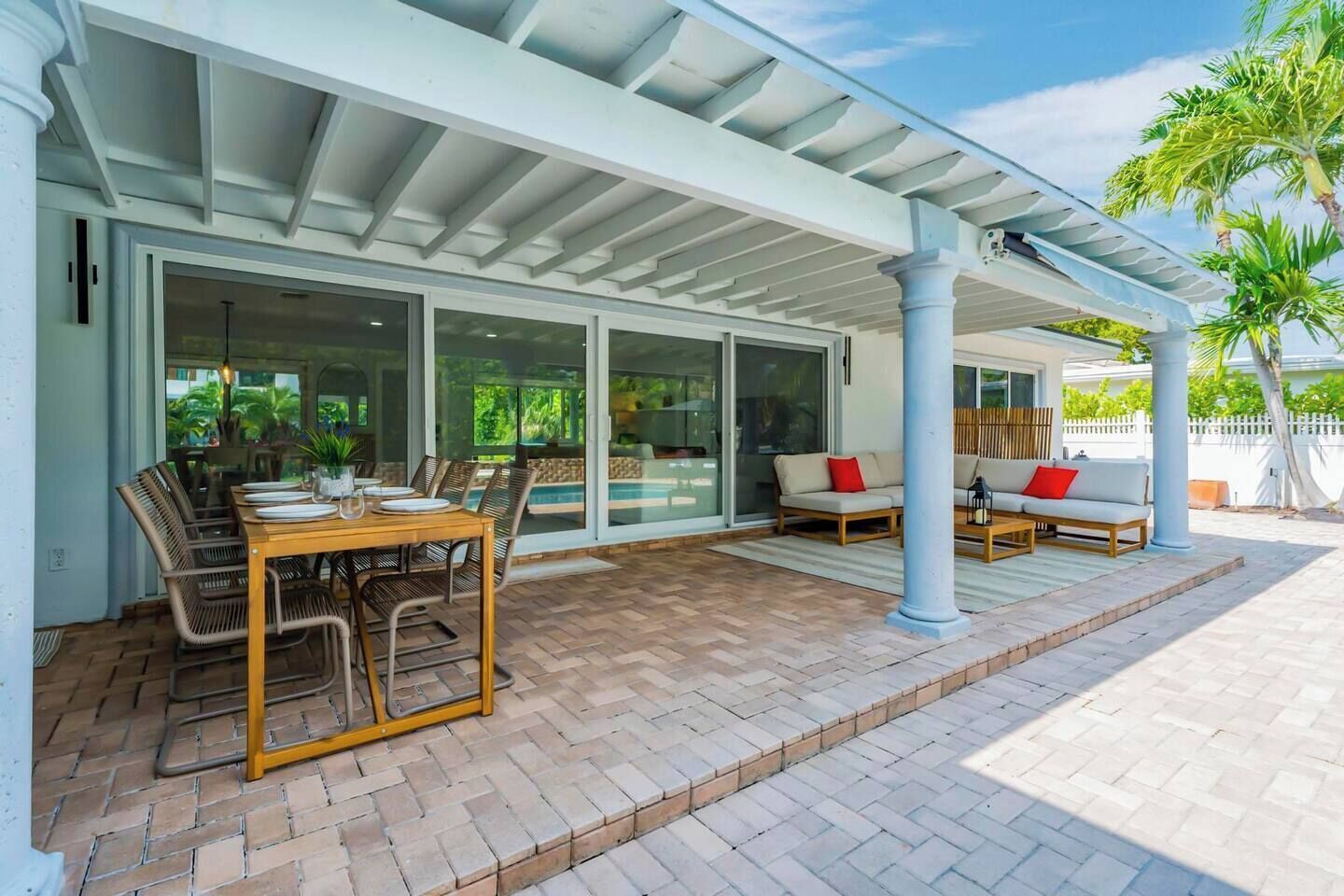 An outdoor patio with a covered dining area, a pool, and a seating area with a white fence and palm trees.