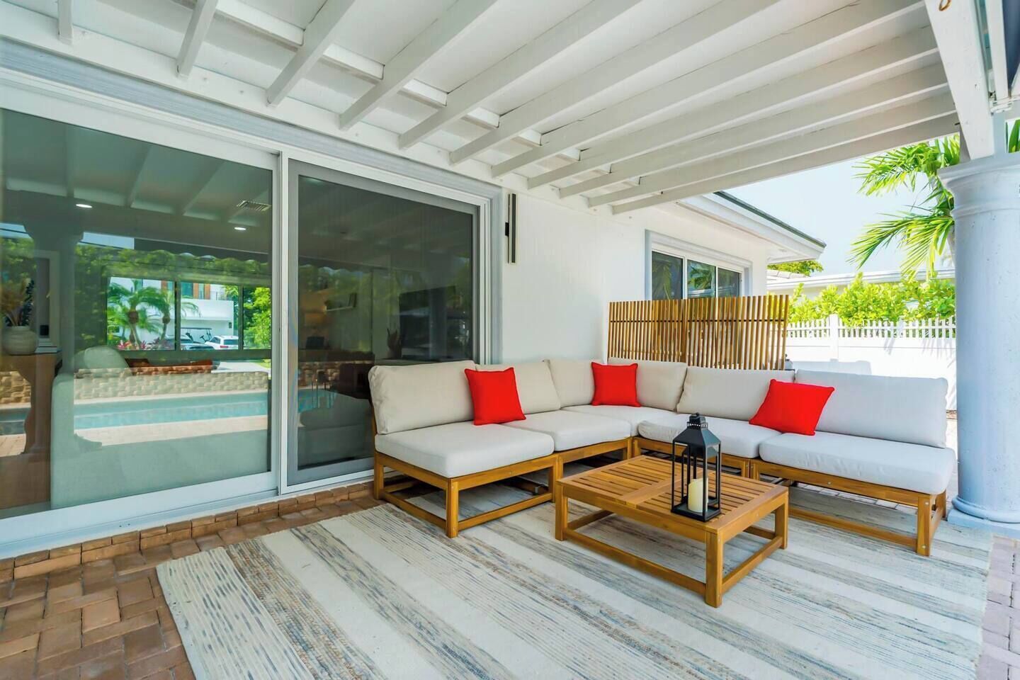 A modern outdoor seating area with a sofa and a coffee table, a glass door leading to a pool, and a wooden fence in the background.
