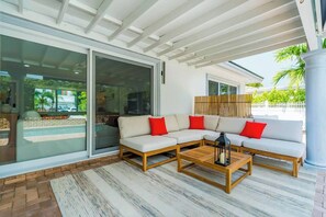 A modern outdoor seating area with a sofa and a coffee table, a glass door leading to a pool, and a wooden fence in the background. - Villa Capucine Beach Retreat | Pool | Games (Pompano Beach)