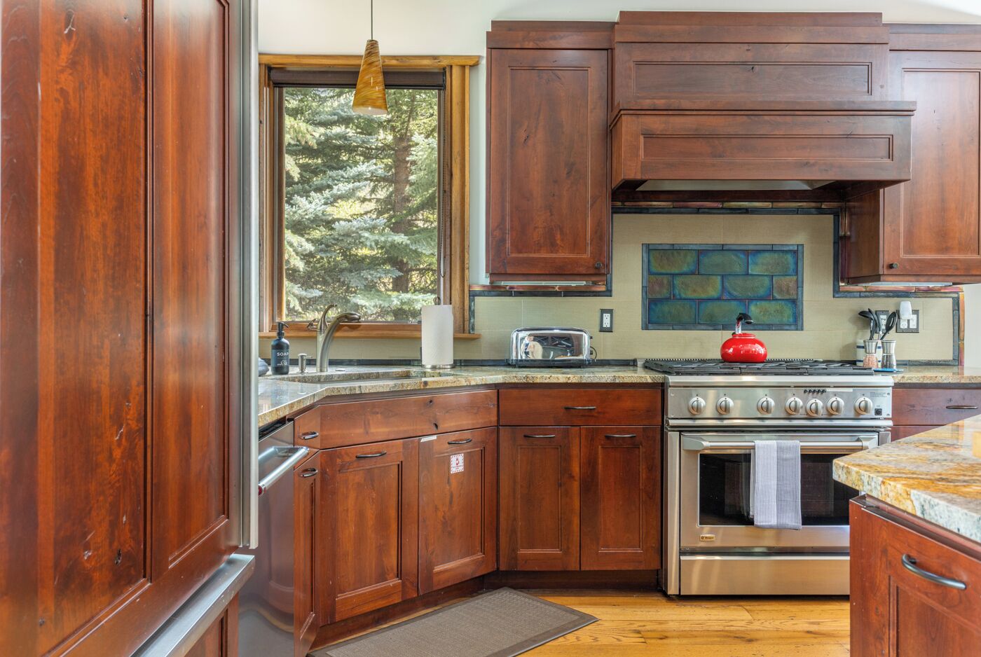 A kitchen with wooden cabinets, a stainless steel stove, and a window with a view of trees.