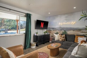 A modern living room with a large TV mounted on the wall, a cozy sofa set, a coffee table, and a view of a mountain landscape through the window. - Hot Tub, Sauna, Fire Pit, Huge Yard near Denver and Red Rocks (Arvada)