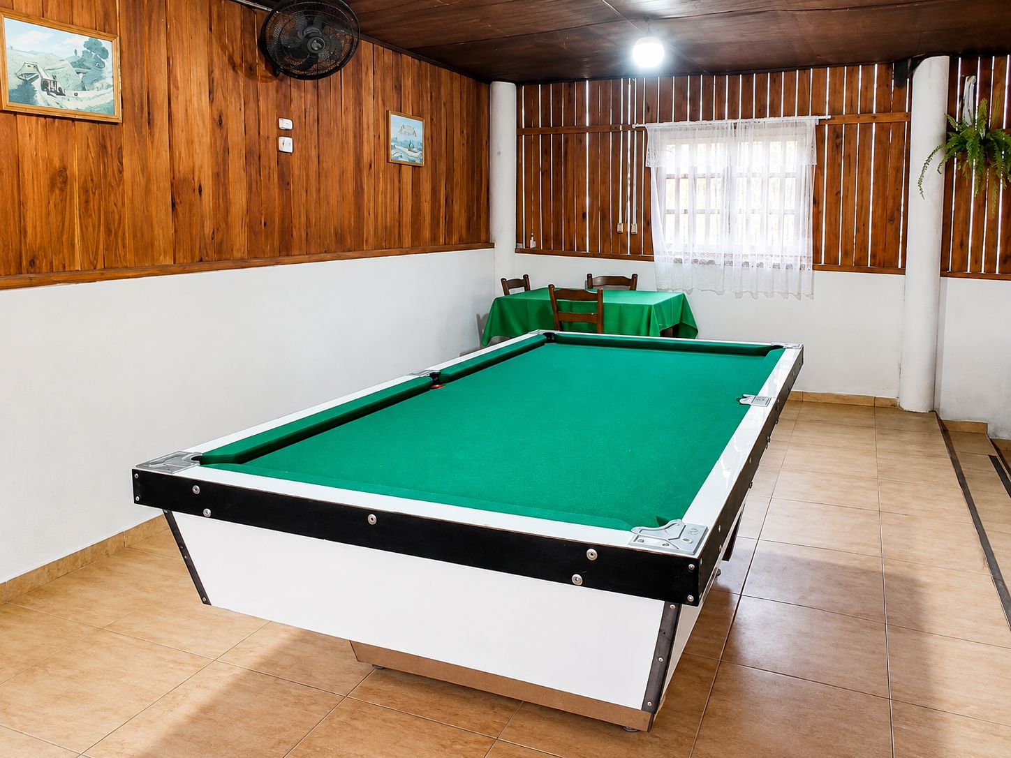 Una mesa de billar en una sala con paneles de madera, un ventilador y una ventana con cortinas.