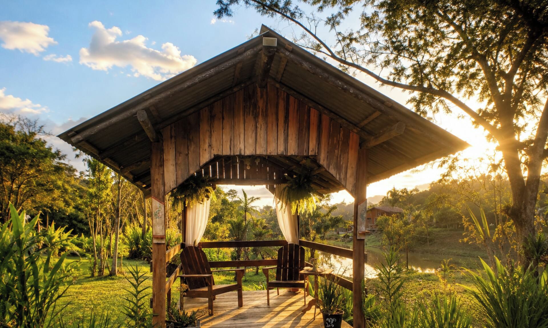 A wooden gazebo with a porch, surrounded by lush greenery and a body of water.