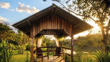 A wooden gazebo with a porch, surrounded by lush greenery and a body of water.
