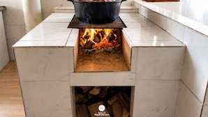 A kitchen with a built-in wood-burning stove, white marble tiles, and a window with wooden blinds.