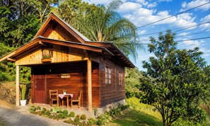 Una cabana de fusta amb un porxo, una taula i cadires, envoltada de vegetació exuberant.