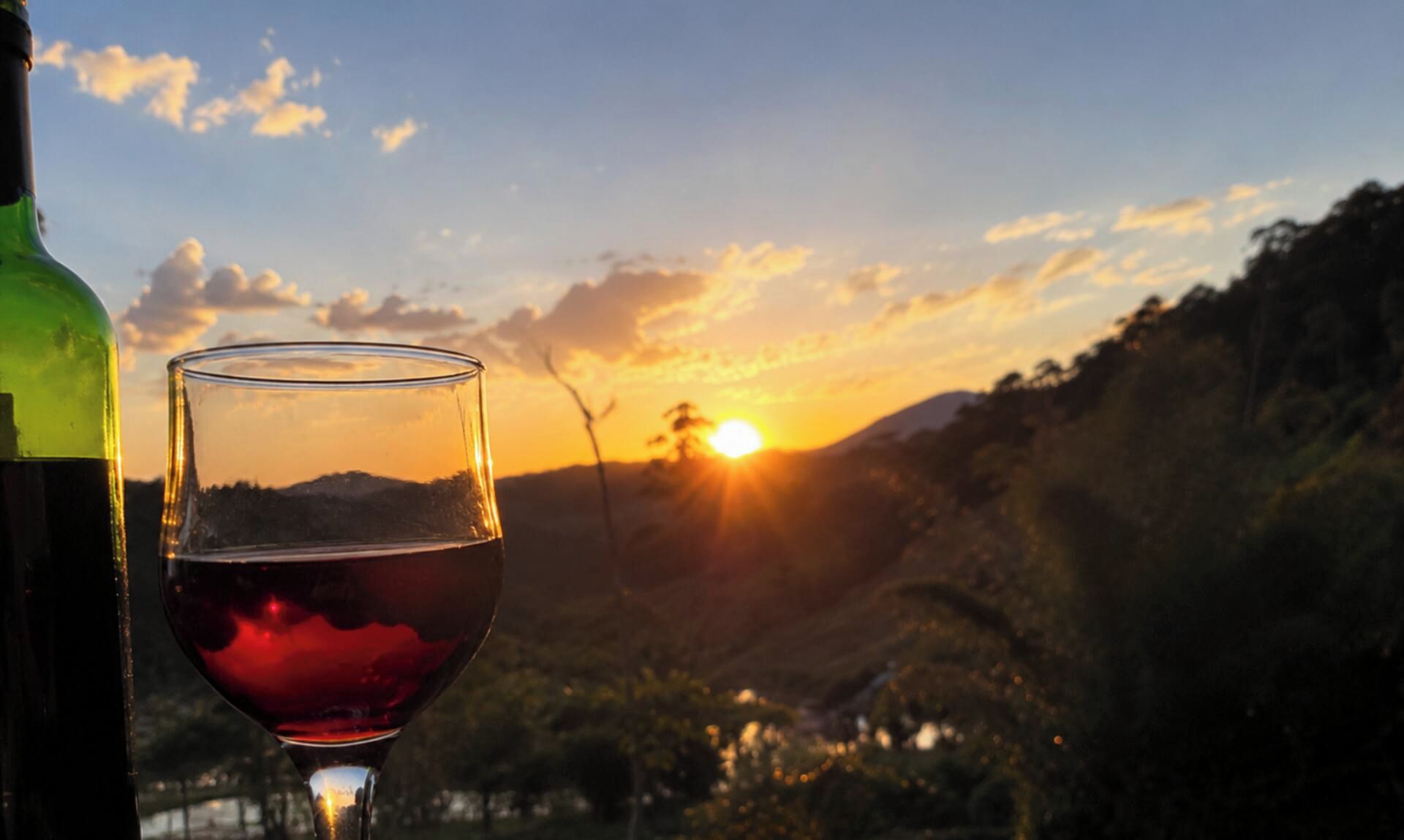 Ein Weinglas mit Rotwein und eine Flasche, im Hintergrund ein Sonnenuntergang und Berge.