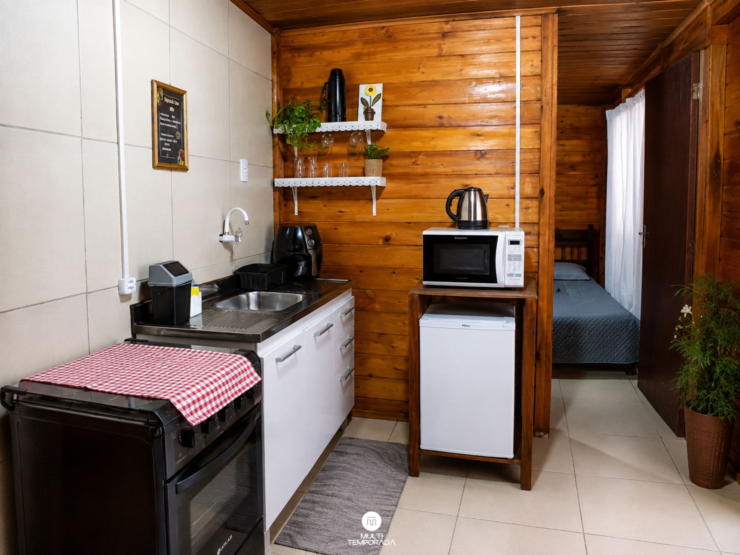 A compact kitchen with a sink, stove, microwave, and a small refrigerator. There is a wooden shelf with plants and a kettle. A bed is visible in the background.
