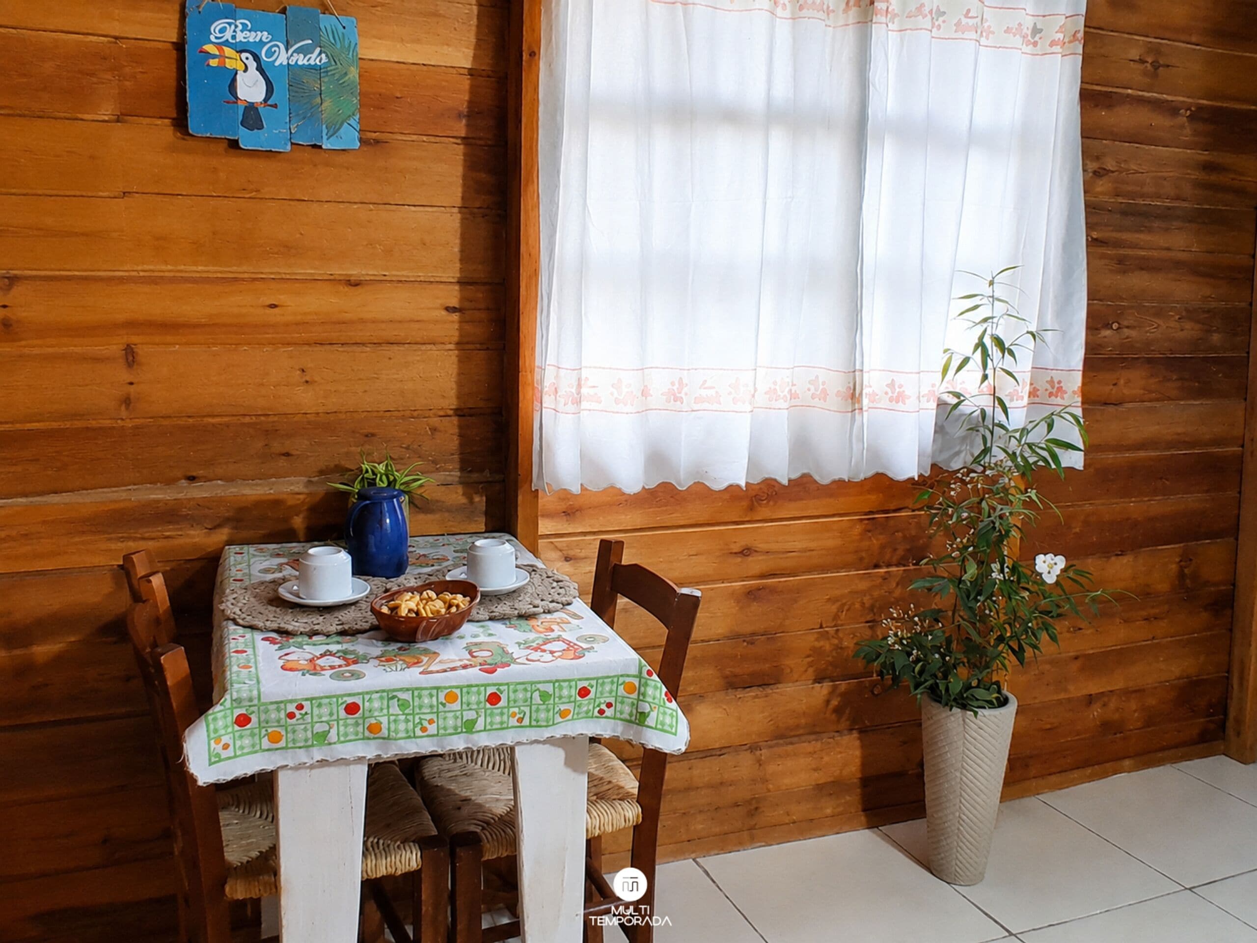 A wooden cabin interior with a dining table set for two, a blue wall decoration, and a window with a white curtain.