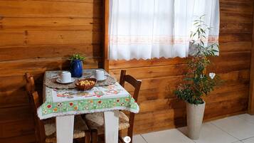 A wooden cabin interior with a dining table set for two, a blue wall decoration, and a window with a white curtain.