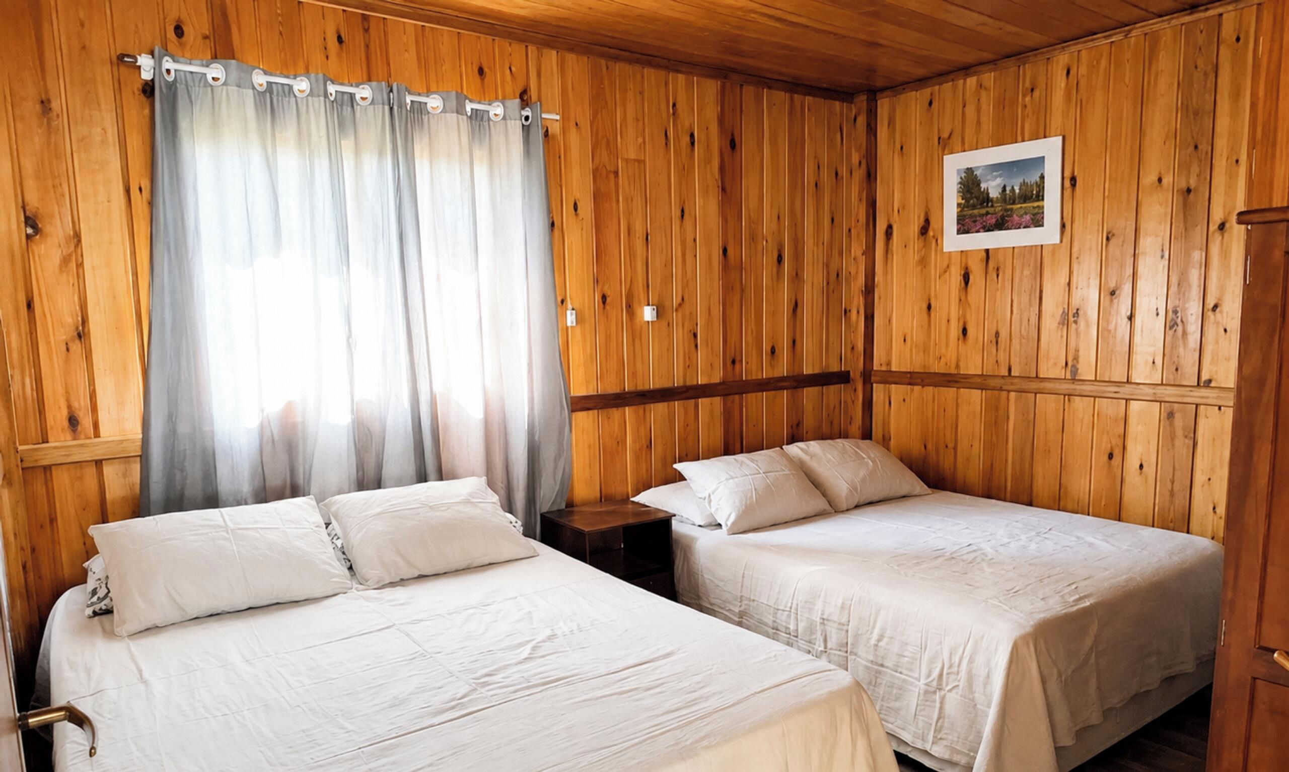 Una habitación con dos camas, paredes de madera, una ventana con cortinas, una mesita de noche y un cuadro en la pared.