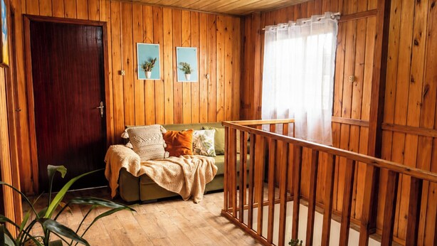 A wooden cabin interior with a wooden door, a sofa with cushions, a window with sheer curtains, and a wooden railing.