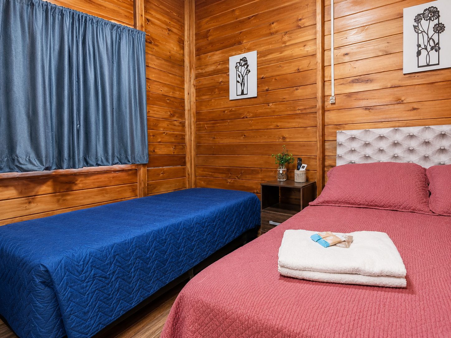 A room with two beds, wooden walls, a window with curtains, and two framed pictures.