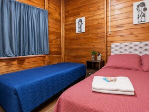 Una habitación con dos camas, paredes de madera, una ventana con cortinas y dos cuadros enmarcados.