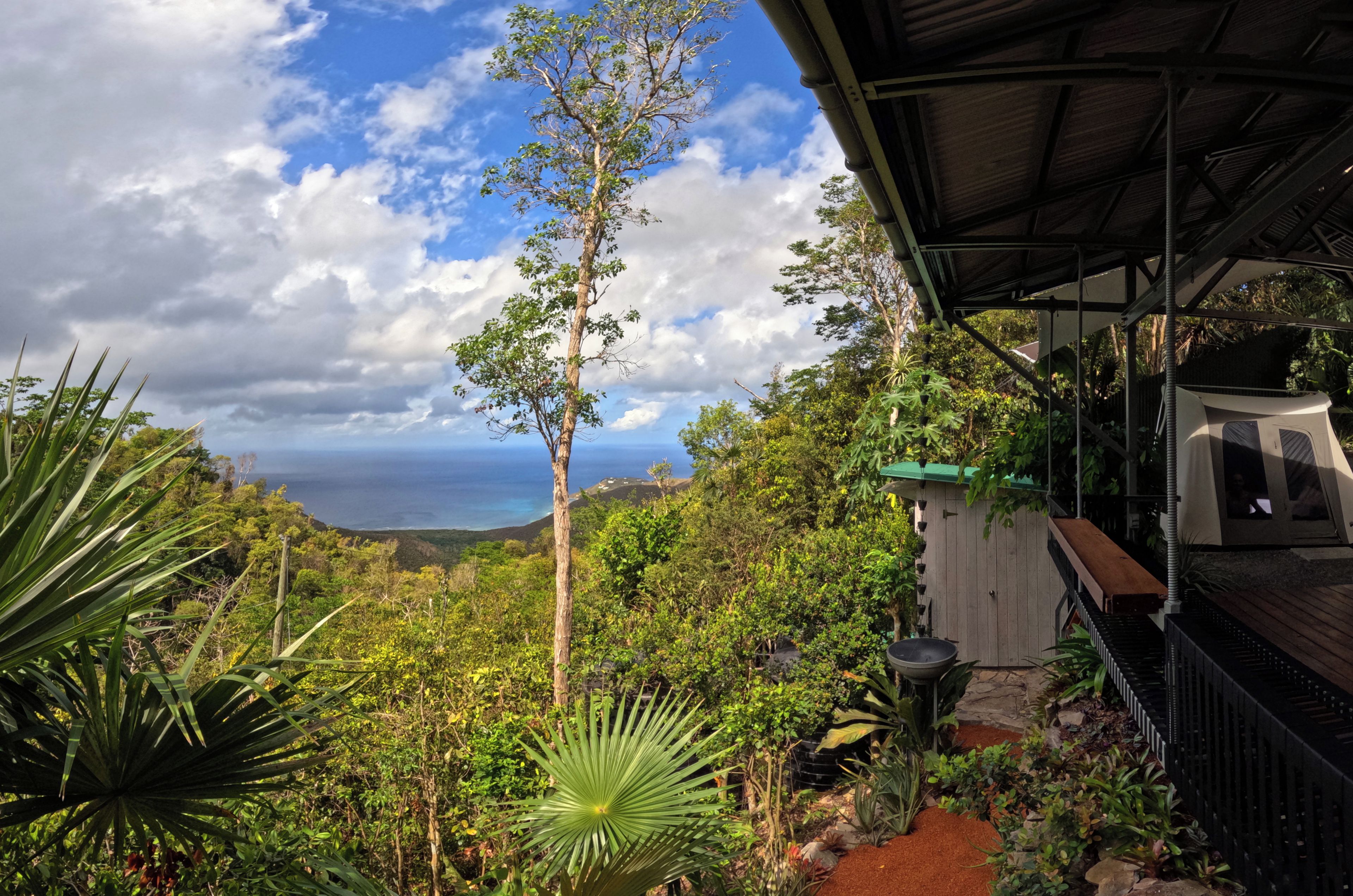 A tropical garden with a view of the ocean, a wooden deck, and a covered seating area.